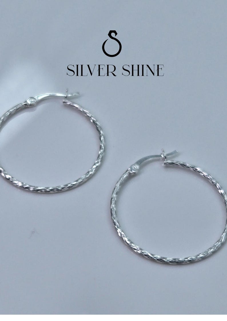 Op de afbeelding: Twee zilveren oorringen met een getextureerd, gedraaid ontwerp. De oorbellen worden gepresenteerd op een lichtgrijze achtergrond. De tekst "SILVER SHINE" staat boven de oorbellen.