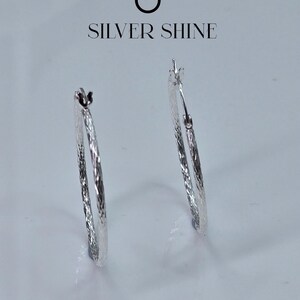 Op de afbeelding: Twee zilveren oorringen met een getextureerd oppervlak worden getoond tegen een witte achtergrond. De oorbellen zijn van het merk "SILVER SHINE". Een klassiek sieraad.