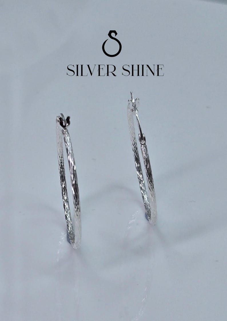 Op de afbeelding: Twee zilveren oorringen met een getextureerd oppervlak worden getoond tegen een witte achtergrond. De oorbellen zijn van het merk "SILVER SHINE". Een klassiek sieraad.