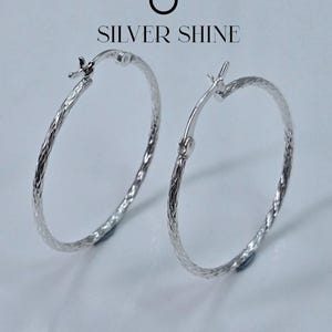 Op de afbeelding: Twee zilveren oorringen met een getextureerd, geweven ontwerp. De oorbellen worden gepresenteerd op een witte ondergrond, met de tekst "SILVER SHINE" erboven. De oorbellen hebben een eenvoudig, elegant ontwerp.