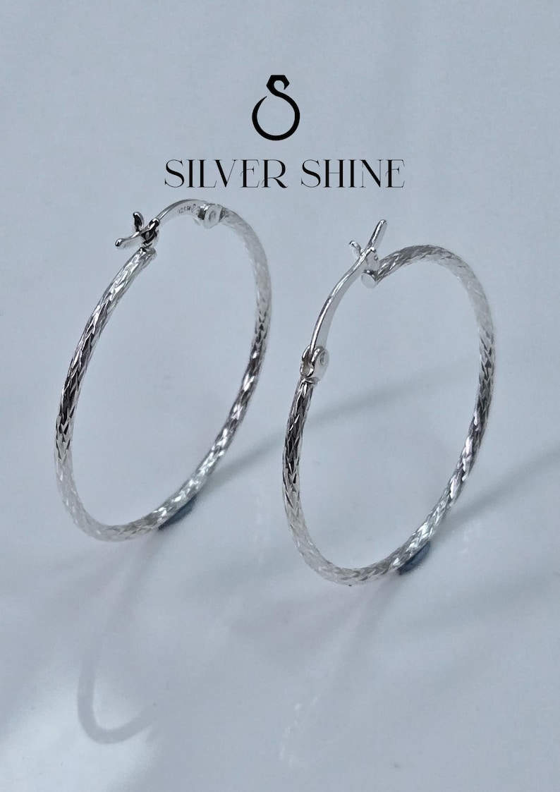 Op de afbeelding: Twee zilveren oorringen met een getextureerd, geweven ontwerp. De oorbellen worden gepresenteerd op een witte ondergrond, met de tekst "SILVER SHINE" erboven. De oorbellen hebben een eenvoudig, elegant ontwerp.