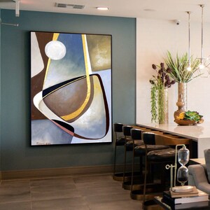 Puede incluir: Pintura abstracta con formas geométricas en tonos de marrón, dorado, azul y blanco. La pintura está enmarcada en negro y cuelga de una pared azul verdosa.