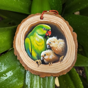 Könnte beinhalten: Handbemalter Holzschmuck mit einem grünen Papagei mit rotem Schnabel und zwei gelben Küken. Das Kunstwerk befindet sich in einer kreisförmigen Holzscheibe mit einer roten und goldenen Schnur zum Aufhängen. Der Hintergrund ist ein üppiges Grün.