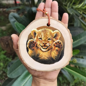May include: A hand-painted wooden ornament featuring a smiling lion cub. The cub is painted in shades of brown and gold, with its paws outstretched. The ornament is round, with a natural wood grain edge, and has a red string for hanging.