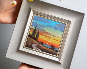 Op de afbeelding: Een kleine ingelijste schildering die een zonsondergang boven een landschap met een weg en bomen afbeeldt. De schildering heeft een zilveren lijst en wordt door twee handen vastgehouden. De lucht gaat over van blauw naar oranje en geel.