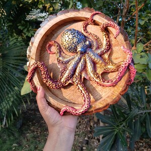 Puede incluir: Una pieza de arte mural de madera con una detallada escultura de pulpo tridimensional. El pulpo está pintado con tonos metálicos dorados, azules y rojos, y está montado sobre una base redonda de madera con un borde natural. La obra de arte se sostiene en una mano.