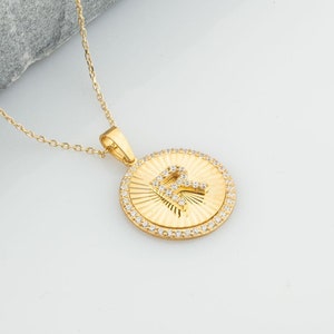 Op de afbeelding: Een gouden ketting met een ronde hanger met de letter "R" omringd door kleine heldere stenen. De hanger heeft een getextureerd ontwerp.