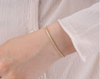 Può includere: Un braccialetto a catena a serpente color oro è indossato al polso di una persona. Il braccialetto ha un design liscio e flessibile. La persona indossa una camicia a maniche lunghe di colore chiaro.