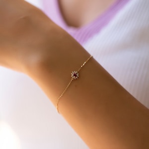 Op de afbeelding: Een delicate gouden kettingarmband met een klein rood edelsteentje omringd door heldere, glinsterende stenen in de vorm van een bloem.