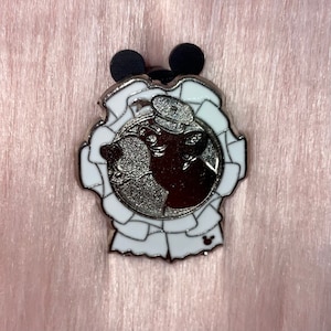 May include: A silver pin with a white ribbon design. The center of the pin features a black and white image of two cartoon characters wearing sailor hats.