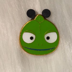 May include: A round metal enamel pin featuring a bright green chameleon face with large white eyes, black pupils, and a thin dark blue line for a mouth. The pin has a gold-colored metal border and two black circular Mickey Mouse ear-shaped backings.