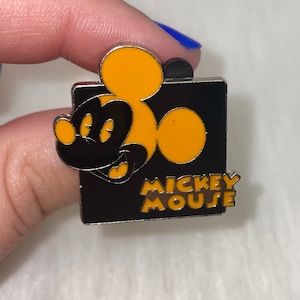 May include: A black and yellow enamel pin featuring Mickey Mouse's head with the text "Mickey Mouse" below.