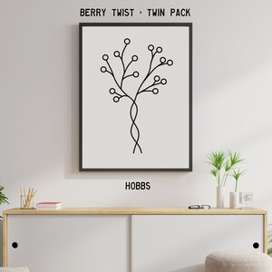 May include: A framed print with a minimalist black and white design of a stylized tree with berries. The print is labeled "BERRY TWIST • TWIN PACK" and "HOBBS". The artwork hangs above a white cabinet with wooden accents.