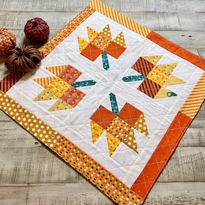 May include: A quilted table runner with a white center featuring four stylized orange and yellow leaves. The border is orange with polka dots and stripes. Decorative pumpkins are in the background.