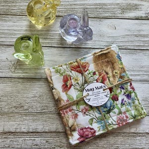 May include: A set of four floral print mug mats tied with twine, featuring a variety of flowers and a bunny design. Three glass bunny candle holders in yellow, clear, and green are also visible.