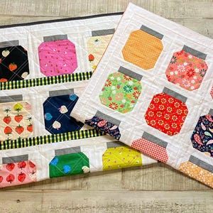 May include: Two rectangular fabric quilts featuring a repeating pattern of colorful mason jars. Each jar displays various floral, strawberry, and geometric prints in shades of red, blue, pink, yellow, and green, finished with grey lids and white borders.