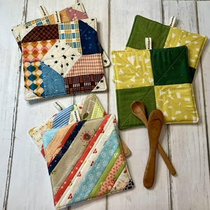 May include: A collection of colorful, quilted pot holders with various geometric patterns. The pot holders feature a mix of fabrics in shades of blue, green, yellow, and red. Two small wooden spoons are also present. Each pot holder has a small tag that says "KarenLee".