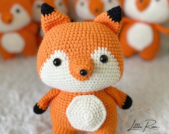 May include: A crocheted orange fox plush toy with black eyes and a white belly. The fox is standing on a white surface.