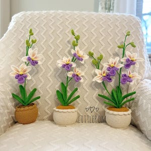 May include: Three crocheted orchid plants in white pots. The orchids have white petals with purple centers and green leaves. The pots are white with a brown rim. The plants are sitting on a white couch with a chevron pattern.