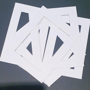 May include: A collection of white picture frame mounts with square apertures. The mounts are made of thick, white card and are arranged in a slightly overlapping pattern. The background is a dark grey surface.