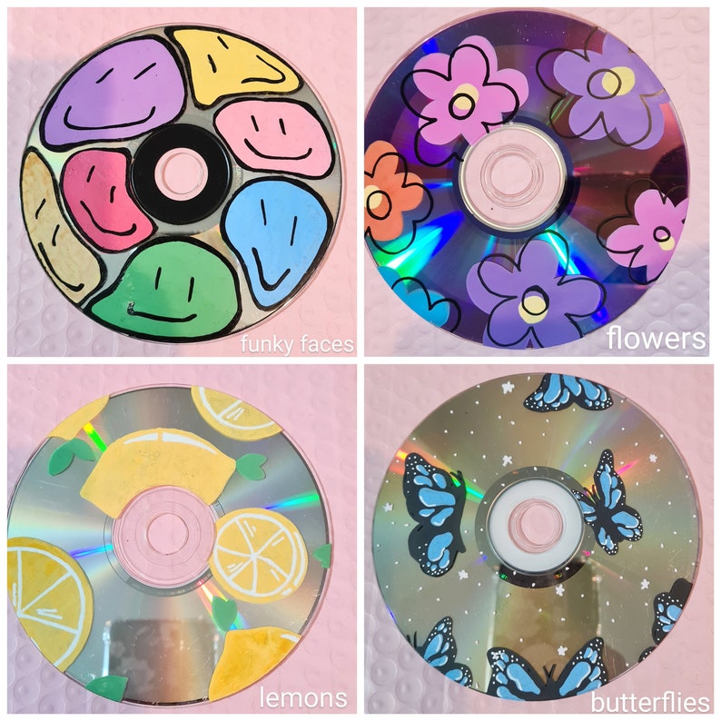 Op de afbeelding: Vier handgeschilderde cd's met verschillende ontwerpen. E&eacute;n heeft kleurrijke smiley-gezichten, een andere heeft paarse en oranje bloemen, een derde toont citroenen en de laatste heeft blauwe vlinders. Elke cd heeft tekstlabels: "funky faces", "flowers", "lemons" en "butterflies".