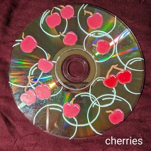Op de afbeelding: Een beschilderde cd met een kersendesign. De kersen zijn rood met groene steeltjes en zijn op de cd gerangschikt met witte luslijnen. Het woord "cherries" staat in het wit onderaan de cd.