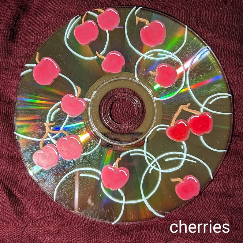 Op de afbeelding: Een beschilderde cd met een kersendesign. De kersen zijn rood met groene steeltjes en zijn op de cd gerangschikt met witte luslijnen. Het woord "cherries" staat in het wit onderaan de cd.