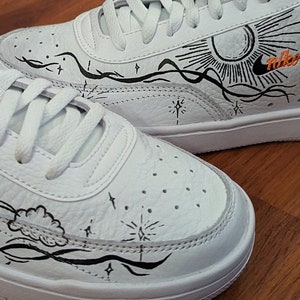 Puede incluir: Zapatillas de cuero blancas con detalles en negro y naranja. Los zapatos presentan ilustraciones dibujadas a mano de un sol, nubes y estrellas. El logo de Nike en naranja es visible en el lateral.