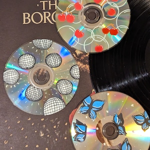 Op de afbeelding: Drie handgeschilderde cd's met unieke ontwerpen. E&eacute;n cd toont rode kersen, een andere heeft zwart-witte discoballen en de derde blauwe vlinders. De cd's zijn gerangschikt op een donkere achtergrond, mogelijk een platenhoes.