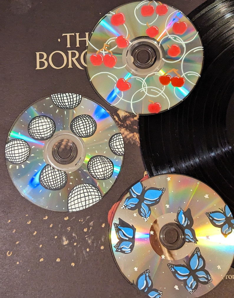 Op de afbeelding: Drie handgeschilderde cd's met unieke ontwerpen. E&eacute;n cd toont rode kersen, een andere heeft zwart-witte discoballen en de derde blauwe vlinders. De cd's zijn gerangschikt op een donkere achtergrond, mogelijk een platenhoes.