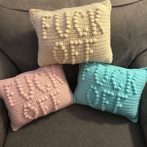 May include: Three crocheted throw pillows with the word "Mum" spelled out in raised stitches. The pillows are beige, pink, and light blue.