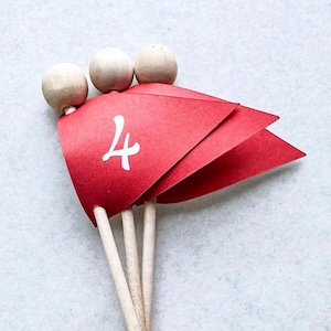 May include: Red flag-shaped party picks with the number 4 in white. Each pick has a wooden stick and a small wooden ball on top. The picks are arranged in a fan shape, perfect for decorating cupcakes or other treats.