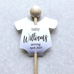 May include: A white baby onesie cutout with a wooden stick and a round wooden topper. The onesie has the text "baby Williams arriving April 2025" printed on it.