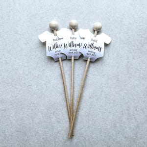 May include: Three white baby onesie shaped paper cutouts on wooden sticks. Each cutout has the text "baby Williams arriving April 2025".