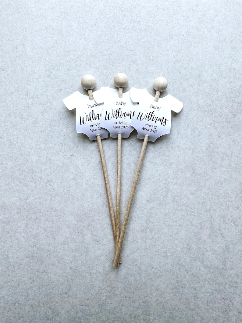 May include: Three white baby onesie shaped paper cutouts on wooden sticks. Each cutout has the text "baby Williams arriving April 2025".
