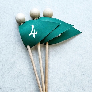 May include: A close-up of four green flag-shaped table decorations. Each flag has a white number printed on it, with the number 4 visible on the front-most flag. The flags are attached to wooden sticks and topped with small wooden spheres.
