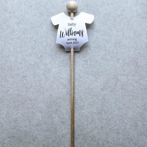 May include: A white baby onesie cutout with the text "baby Williams arriving April 2025" on a wooden stick.