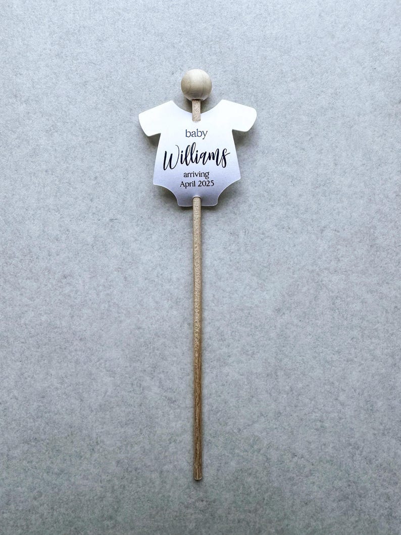 May include: A white baby onesie cutout with the text "baby Williams arriving April 2025" on a wooden stick.