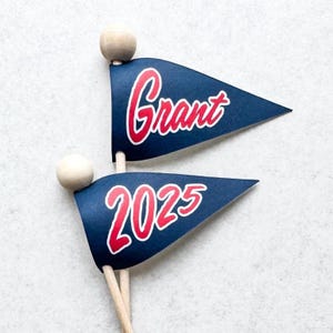 May include: Two blue pennant flags with wooden sticks. The top flag has the name "Grant" written in red cursive. The bottom flag has the year "2025" written in red cursive.