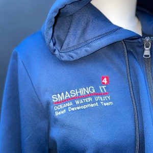 May include: A navy blue zip-up hoodie with a red embroidered logo that reads "Smashing It 4 Oceania Water Utility Sales Development Team".