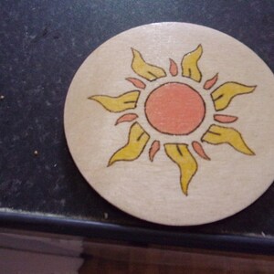 May include: A wooden coaster with a hand-painted sun design. The sun is yellow with orange rays and a pink center.