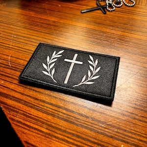 May include: A rectangular black embroidered patch featuring a white cross centred between two white laurel leaf branches. The patch has a stitched border and is positioned on a surface with a wood grain pattern, near a silver chain and cross.