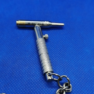 May include: A silver metal keychain tool with a T-shaped handle and a small, pointed tip. The tool has a textured grip and a small knob on the side. The keychain is attached to a silver chain.
