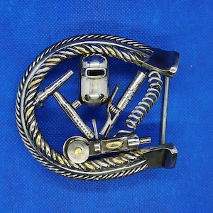 May include: A silver metal belt buckle shaped like a horseshoe with a welding mask and various tools attached to it.