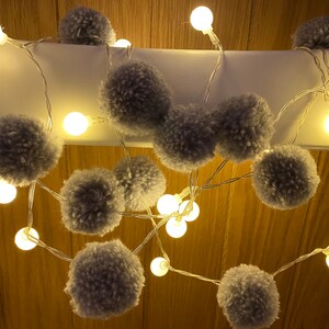 May include: A string of white fairy lights with gray pom poms hanging from them. The lights are strung across a wooden surface.