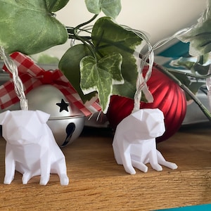 May include: White geometric pug-shaped string lights. The lights are strung on a clear wire and are placed near a red ornament and a silver bell with a star cutout. Green leaves and a red and white checkered bow are also visible.
