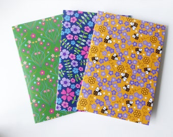 May include: Three notebooks with different floral patterns. The first notebook has a green cover with pink and white flowers and green leaves. The second notebook has a blue cover with pink, white, and blue flowers and green leaves. The third notebook has a yellow cover with purple flowers and bees.