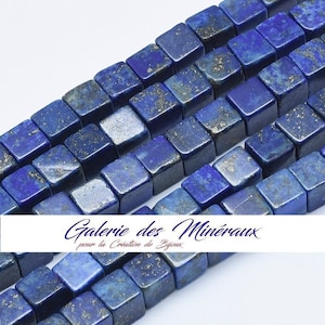 Puede incluir: Una hebra de cuentas de lapislázuli azul oscuro, cada cuenta tiene forma de cubo con motas doradas. Las cuentas están dispuestas en filas y están listas para la elaboración de joyas.