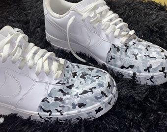 May include: Pair of white Nike Air Force 1 sneakers with a custom camouflage design on the toe box and sides. The camouflage pattern features shades of gray and black. White laces and a silver metal tag complete the look.