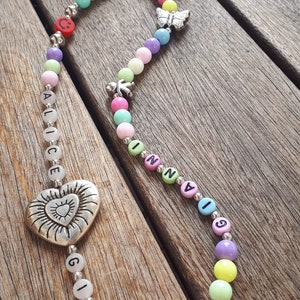 May include: A colorful beaded phone charm with the names Alice, Gio, and Inna spelled out in black letters. The charm also features a silver heart with a sun design, a butterfly, a flower, and a small yellow charm.